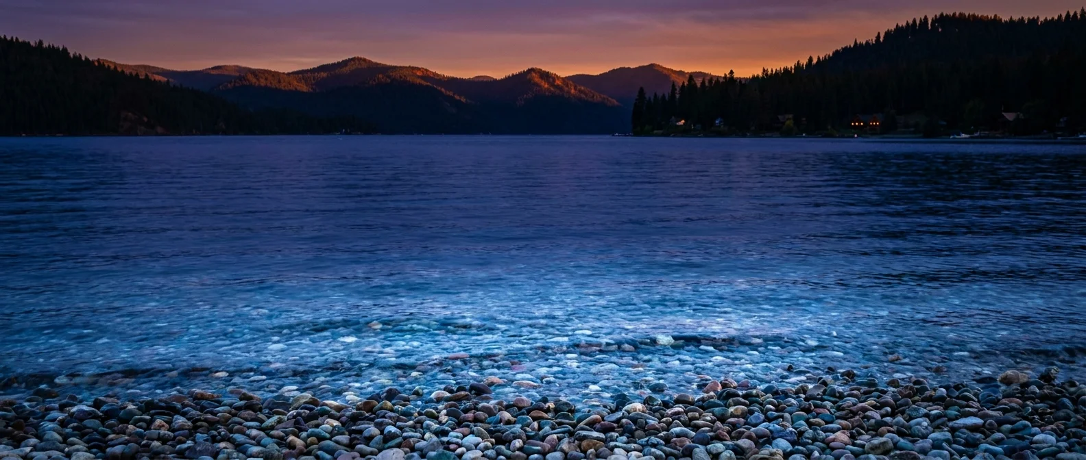 A tranquil evening view of Lake Coeur d'Alene with mountain silhouettes reflected in the water at sunset.