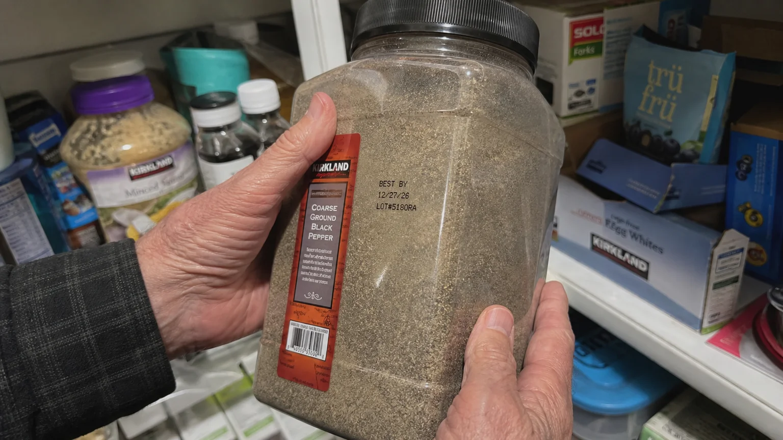 A close-up photo of a senior checking the expiration date on a large bulk container in a crowded pantry.