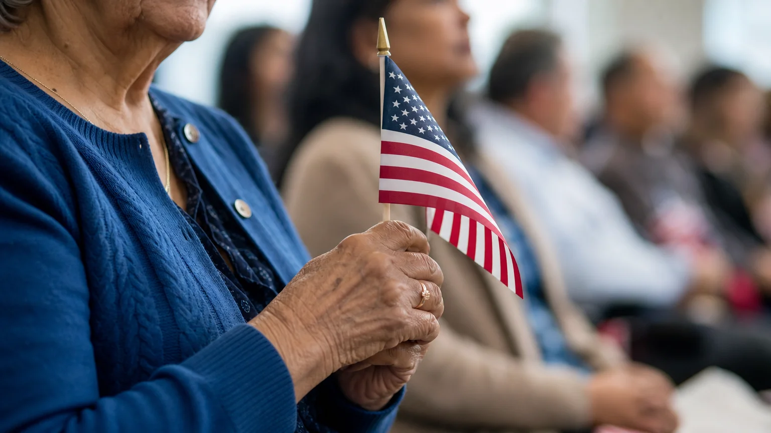 A close-up photograph of an older woman's hands holding a small American flag at a citizenship ceremony.