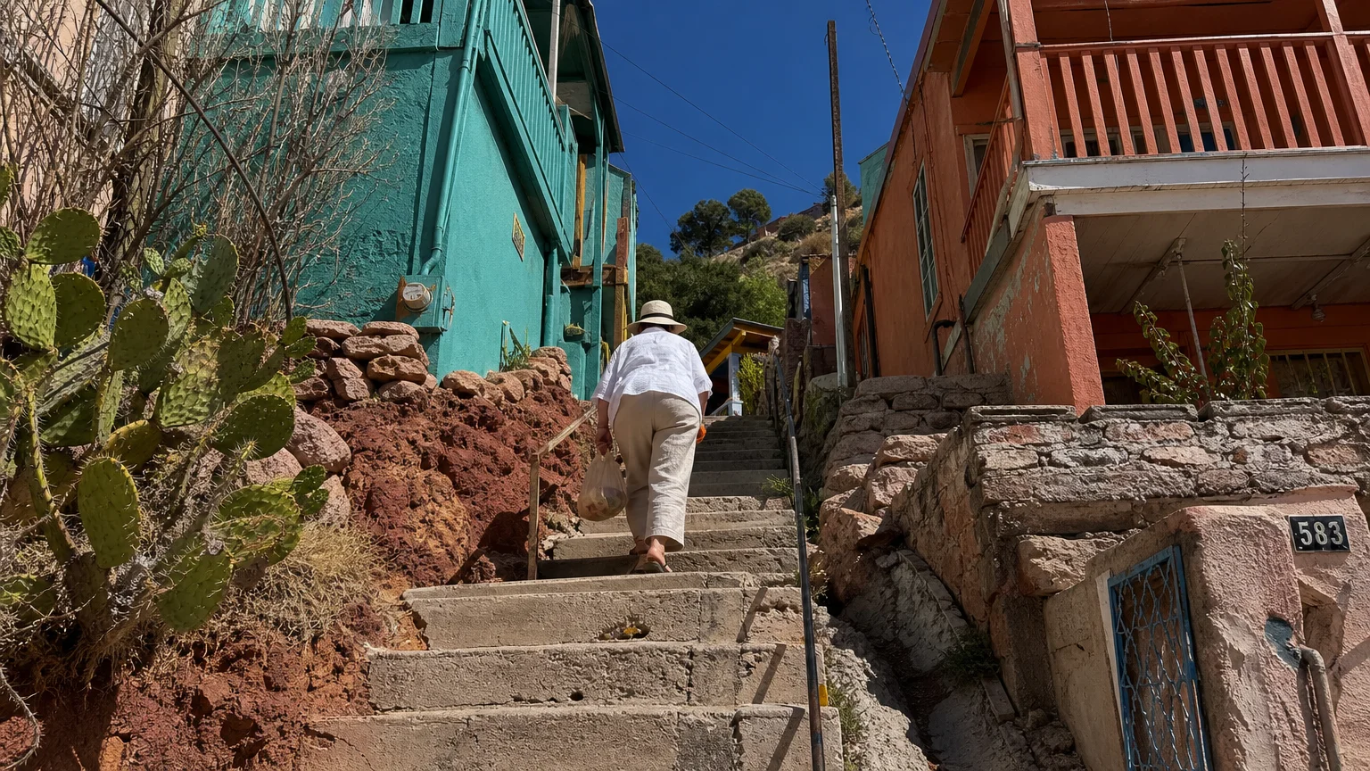 A senior woman carrying groceries walks up a long, steep outdoor staircase between colorful desert houses.