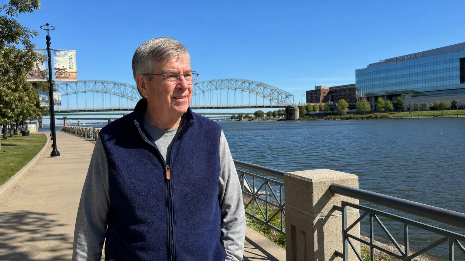 An older man walking along the Peoria riverfront trail with the Illinois River and a modern medical center visible in the background.