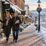 An older couple walks along a cleared brick sidewalk looking into shop windows on a snowy main street.